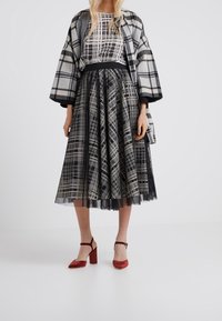 Woman wearing black and white plaid jacket, matching patterned skirt and top, paired with red velvet ankle-strap block heels against white background.