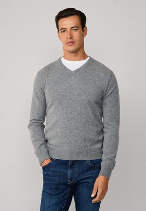 Hombre con jersey gris de cuello en V sobre camisa blanca y vaqueros azules, de pie con una mano en el bolsillo contra un fondo liso.