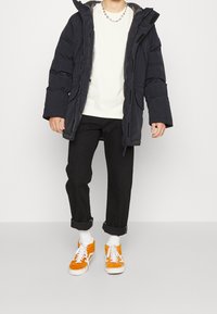 Personne portant une doudoune noire par-dessus une chemise blanche, un pantalon noir retroussé, des chaussettes blanches et des baskets orange vif à lacets blancs.