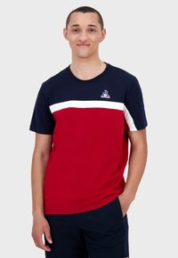 Röd och marinblå kortärmad t-shirt i bomull. Har ett vitt horisontellt rand över bröstet och en logotyp på övre vänstra sidan.