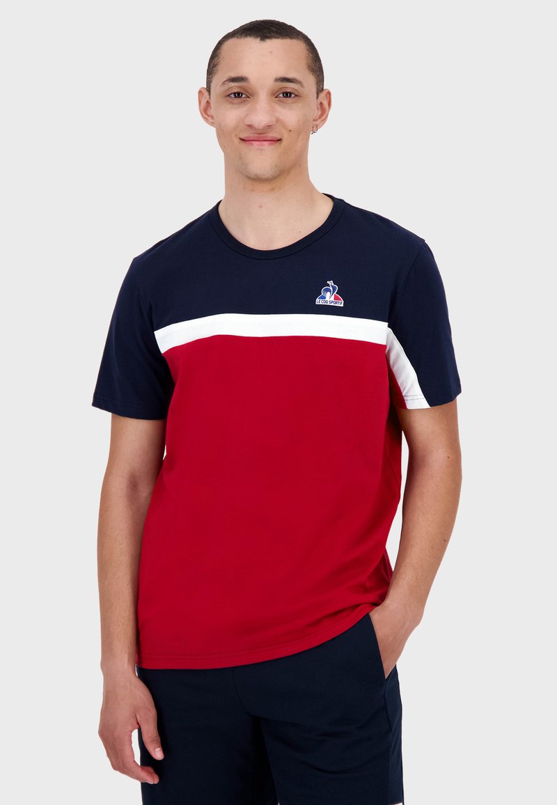 Röd och marinblå kortärmad t-shirt i bomull. Har ett vitt horisontellt rand över bröstet och en logotyp på övre vänstra sidan.