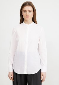 Camicia bianca a maniche lunghe con colletto, realizzata in tessuto leggero, caratterizzata da una linea dritta e un orlo curvo.