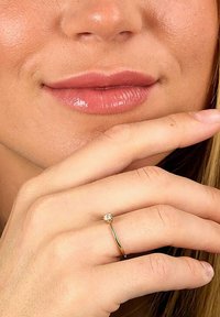 Goldring mit einem kleinen klaren Edelstein, getragen an einer Hand mit heller Hautfarbe, die glatte Haut und natürliche Nägel zeigt.