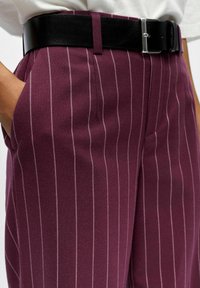 Pantalons bourgogne avec des rayures blanches verticales, dotés de poches latérales et d'une ceinture noire avec une boucle en argent, mettant en avant un tissu texturé.