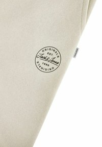 Beige Stoff mit glatter Textur, der ein schwarzes, kreisförmiges Logo von "Jack & Jones Originals" zeigt, das 1990 gegründet wurde.