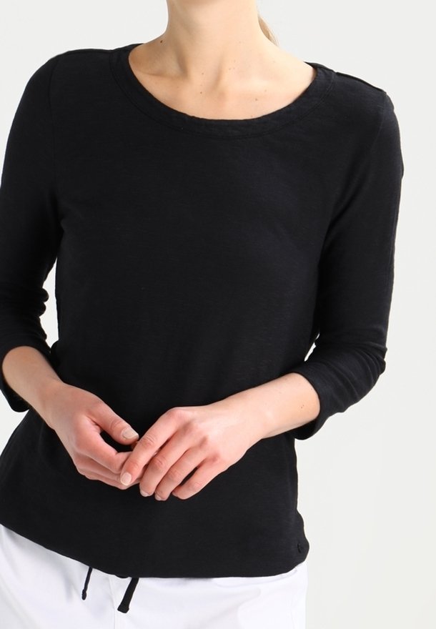 Personne portant un haut noir ajusté à manches longues avec un col rond, mains légèrement jointes, et un pantalon blanc sur un fond uni.