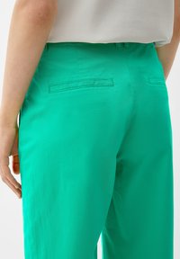 Grüne Baumwollhose mit glatter Textur, zwei Gesäßtaschen und geradem Bein, seitlich betrachtet.