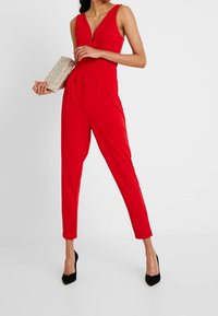 Mono rojo con escote en V profundo, silueta ajustada y piernas afinadas. Combinado con un bolso brillante y tacones puntiagudos negros.