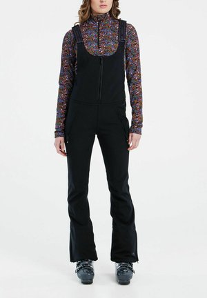 Black ski overall with shoulder straps, zippered front, and zippered pockets. Worn over a multicolored printed long-sleeve top.