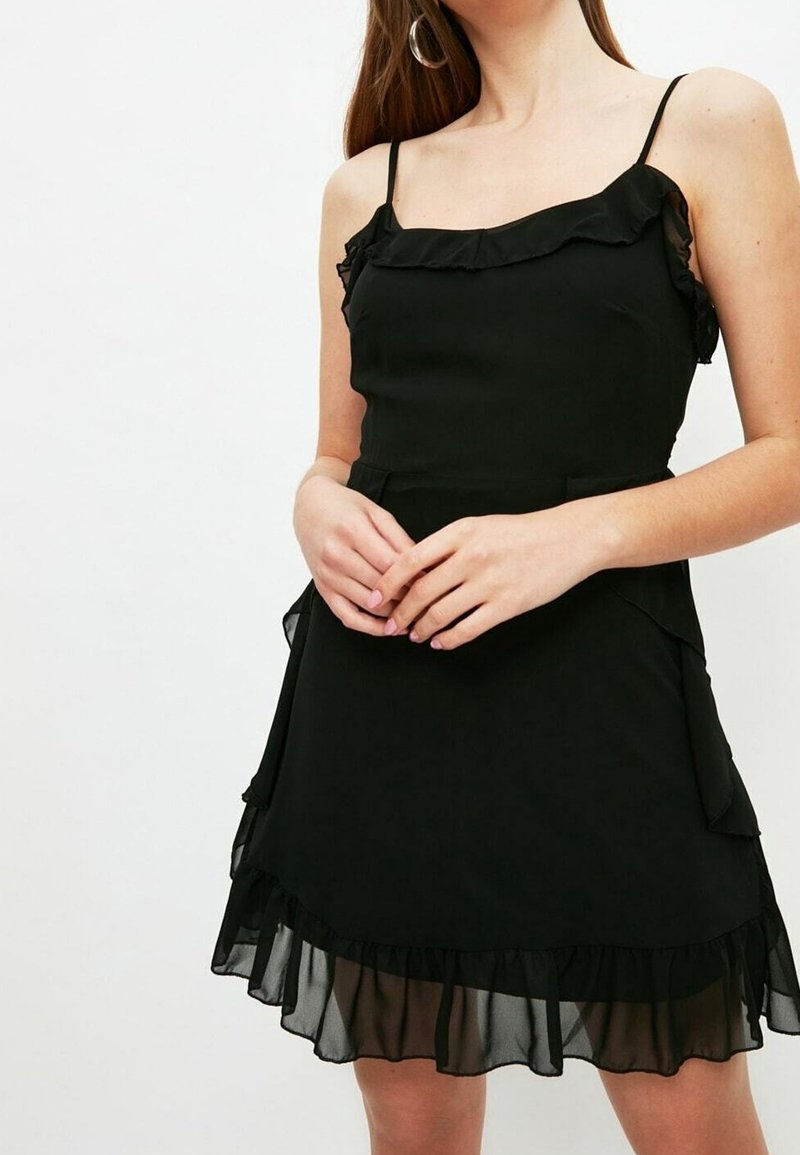 Femme portant une robe noire sans manches avec des volants au niveau de l'encolure et de l'ourlet, debout sur un fond blanc uni.