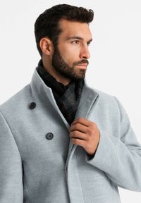 Abrigo de mezcla de lana gris con un cuello alto, dos grandes botones negros y una textura suave; combinado con una bufanda de cuadros oscuros.