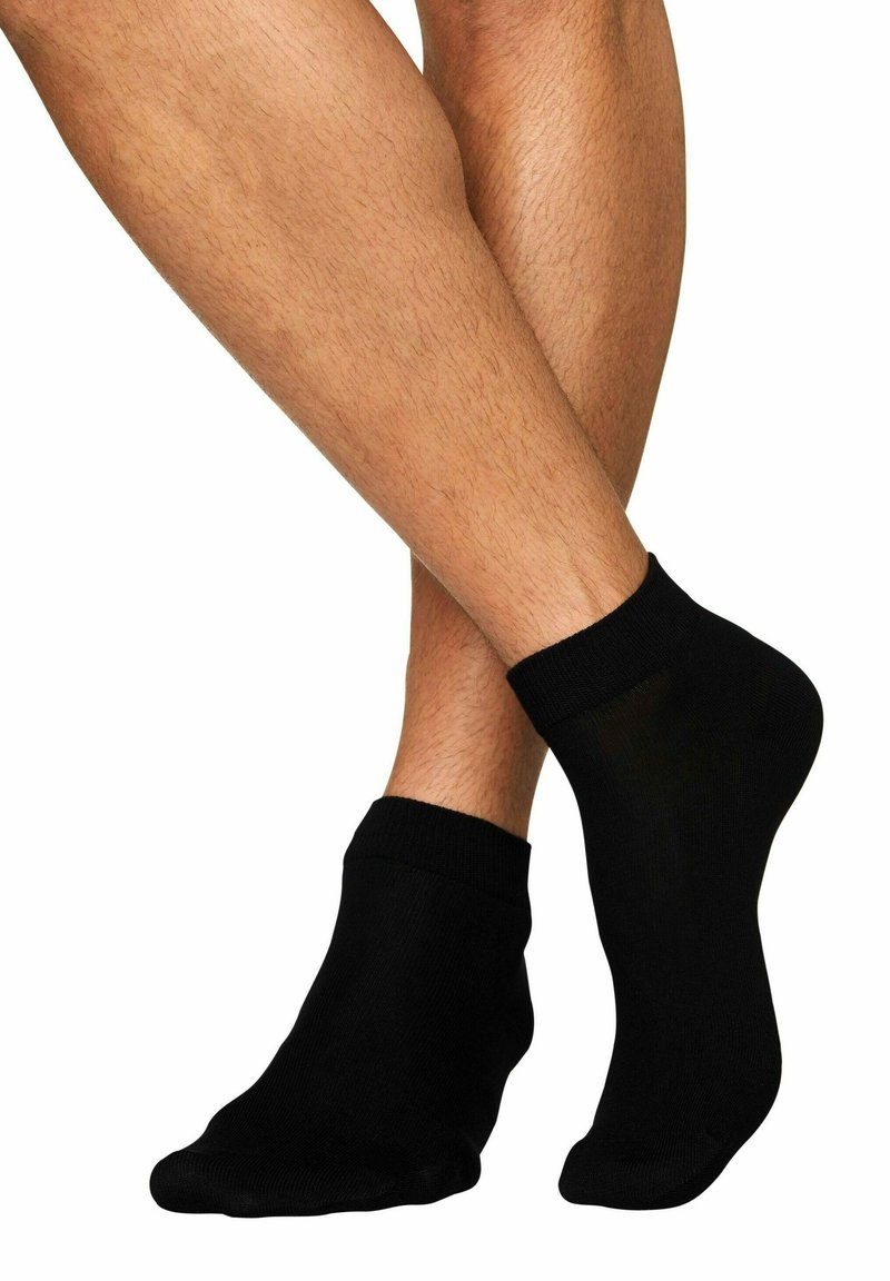 Schwarze Knöchelsocken aus einem glatten, dehnbaren Material, mit einem gerippten Bündchen und einer nahtlosen Zehenpartie. Getragen an übereinandergeschlagenen Beinen.