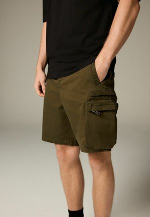 Olive green cargo shorts with multiple pockets, adjustable straps, and a soft, lightweight fabric. Paired with a black t-shirt and dark socks.