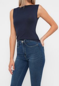 Top sin mangas de color navy hecho de tejido suave, con un cuello alto. Combinado con unos jeans ajustados de talle medio de color azul, con costuras visibles y bolsillo delantero.