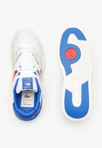 Zapatilla deportiva blanca con acentos en azul y rojo, que cuenta con un superior texturizado, cierre con cordones y una suela de goma con patrón.