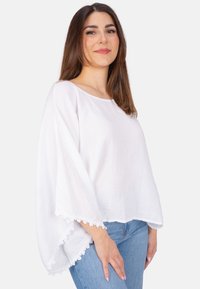 Weiße, leichte Bluse mit lockerem Schnitt und weitem Ärmel. Verfügt über einen strukturierten Stoff und Spitzenbesatz am Saum. Kombiniert mit blauen Jeans.