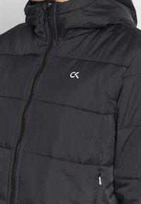 Sort quiltet jakke med lynlås fortil, et lille hvidt "CK" logo på brystet og et nærbillede af den øverste del af torsoen.