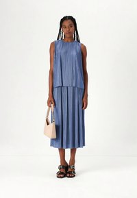 Világoskék pliszírozott ruha ujjatlan felsőrésszel, több réteges kialakítással és folyékony szoknyával. Bézs kézitáskával és fekete szandállal párosítva.