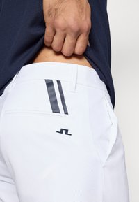 Λευκά παντελόνια με υφή, με σκούρες μπλε ρίγες στην πίσ ω τσέπη και ένα μικρό λογότυπο. Ορατό μέρος από ένα σκούρο μπλε τοπ.