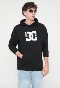Black hoodie with white DC logo and star, made of soft fabric, featuring a kangaroo pocket and drawstring hood, worn with light blue jeans.