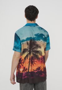 Camicetta a maniche corte con stampa di un tramonto tropicale e palme. I colori variano dal blu all'arancione, con una texture liscia e lucida.