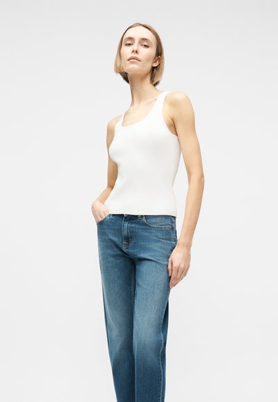 Γυναίκα που φορά λευκό αμάνικο πλεκτό τοπ και μπλε τζιν, στέκεται με το ένα χέρι στην τσέπη, μπροστά σε απλό φόντο.