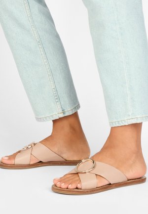 Fødder iført lysebeige slip-on sandaler med brede krydsede remme og stor rund metalspænde, kombineret med lyseblå korte jeans.