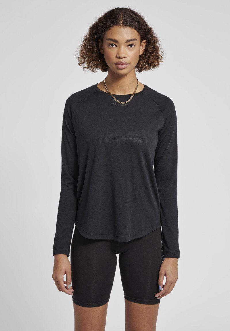 Jonge vrouw met krullend haar, gekleed in een zwarte longsleeve en zwarte fietsbroek, staand voor een effen grijze achtergrond.