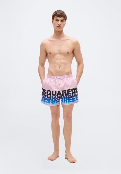 Άνδρας χωρίς πουκάμισο, όρθιος, ξυπόλυτος, φορώντας ροζ μαγιό με μαύρο και μπλε επαναλαμβανόμενο λογότυπο "DSQUARED2", με τα χέρια στις τσέπες, σε απλό φωτεινό φόντο.