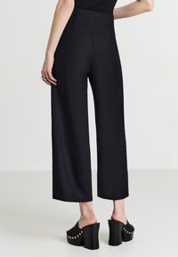 Pantaloni neri a gamba larga con una texture liscia, vita alta e una leggera lucentezza; abbinati a mule nere con plateau e dettagli dorati.