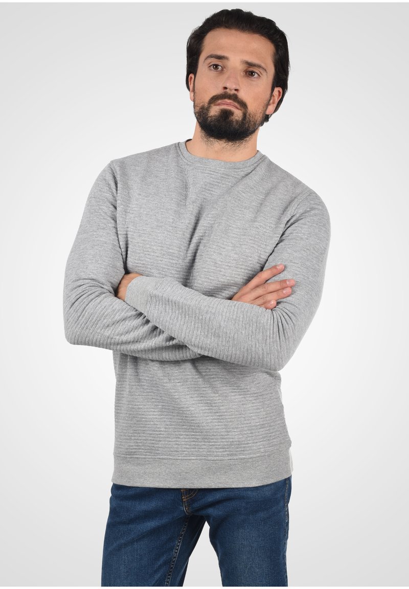 Light gray ribbed sweater with long sleeves, round neckline, and textured design. Worn with blue jeans against a neutral background.