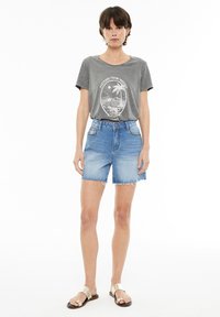 Camiseta de algodón gris con estampado gráfico combinada con pantalones cortos de mezclilla azul claro que presentan un dobladillo deshilachado y una cintura ajustada. Llevada con sandalias doradas.