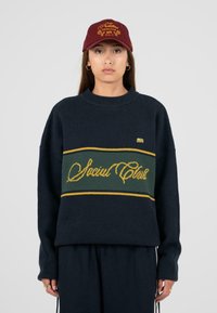 Granatowy sweter o luźnym kroju, z zielono-żółtym poziomym paskiem, haftowanym napisem "Social Club", ryflowanym dekoltem oraz szerokimi rękawami. Burgundowa czapka.