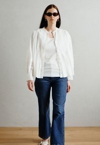 White, textured bomber jacket over a fitted white top, paired with dark blue flared jeans. Model wears dark sunglasses and earrings.