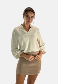 Lichtbeige blouse met een kraag en geplooide mouwen, gecombineerd met een tan, korte naboots-suède rok, wat een gelaagde look creëert.