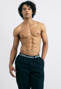 Fekete sportnadrág fehér márkázott derékkal, egy göndör hajú, atlétikus testfelépítésű személy által viselve, sima háttér előtt.