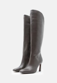 Höga grå läderstövlar med spetsiga tår och höga smala klackar, med sidodragkedjor synliga, stående på en vit yta.