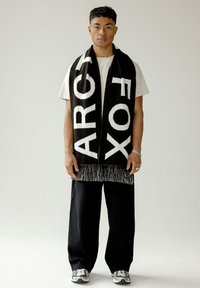 Schwarz-weißer Strickschal mit Fransen, der mit fett gedruckten Buchstaben verziert ist. Kombiniert mit einem cremefarbenen T-Shirt und schwarzen Hosen für den Kontrast. Die Sneaker sind grau.