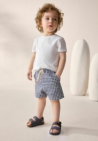 Vit bomullströja, blå och vit rutiga shorts med beige dragsko, grå sandaler med spännen och lockigt hår. Minimalistisk bakgrund.