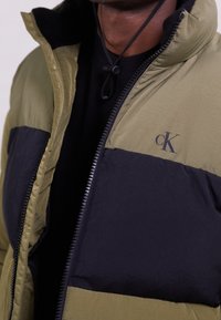 Grön och svart puffjacka med dragkedja, med en slät yta och en logo på bröstet. Justerbara snoddar kring kragen.