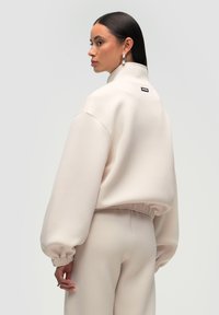 Lys beige fleece sweatshirt med høj krave, elastiske manchetter og cropped nederste kant, med et lille mærke på ryggen. Løs pasform.