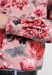 Bloemenblouse van roze satijn met grote rode en zwarte bloemen, grijze mottenprint, knoopmanchet en zwarte taillebanddetail.