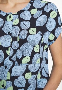 Blusa a maniche corte con sfondo nero, caratterizzata da un motivo a foglie in blu chiaro e verde. Visibili botttoni e colletto rotondo.