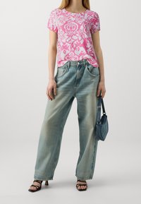 Camisa rosa de manga corta con un estampado floral, combinada con unos vaqueros de pierna ancha en azul claro. La modelo sostiene un pequeño bolso de vaquero y lleva puestas sandalias negras de tacón con tiras.