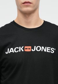 Melna kokvilnas T-kreklā ar balto logotipu un sarkanām akcentiem, kurā rakstīts "JACK" virs "JONES." Klasisks apaļš kakls, mīksta tekstūra.