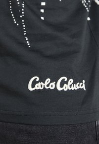 Zwarte T-shirt met een wit pixelpatroon, met een geborduurd "Carlo Colucci" logo aan de onderkant. Katoenen materiaal, gladde textuur.
