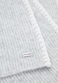 Sciarpa grigia lavorata a maglia con texture a coste, bordi bianchi sfrangiati e piastra in argento con il logo "WITTCHEN" attaccata.
