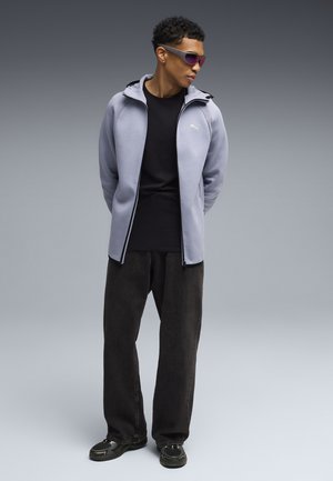 Jeune homme portant des lunettes de sport aux verres teintés violets, un sweat à capuche zippé gris clair, un t-shirt noir, un pantalon noir ample et des baskets, debout les mains derrière le dos.