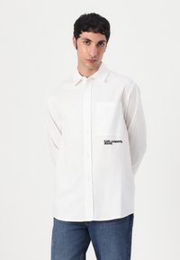 White button-up shirt with a single chest pocket and "KARL LAGERFELD JEANS" embroidered in black on the front. Cotton fabric, relaxed fit.