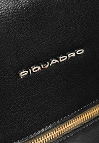 Surface en cuir noir avec un grain texturé, une fermeture éclair dorée et un logo métallique "Piquadro" bien visible.